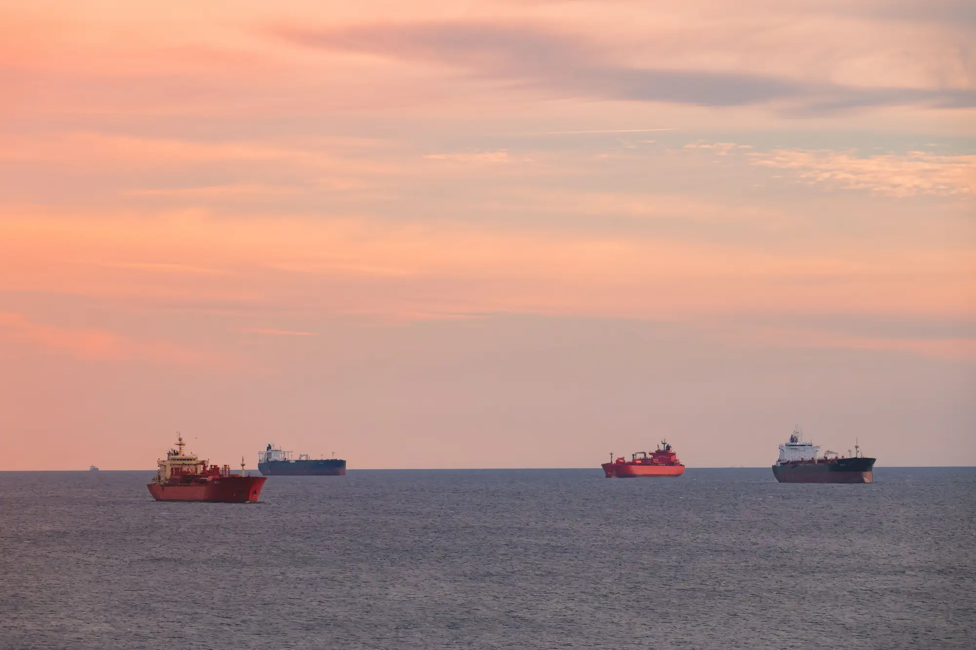 Four cargo ships riding at anchor offshore under a pink evening sky